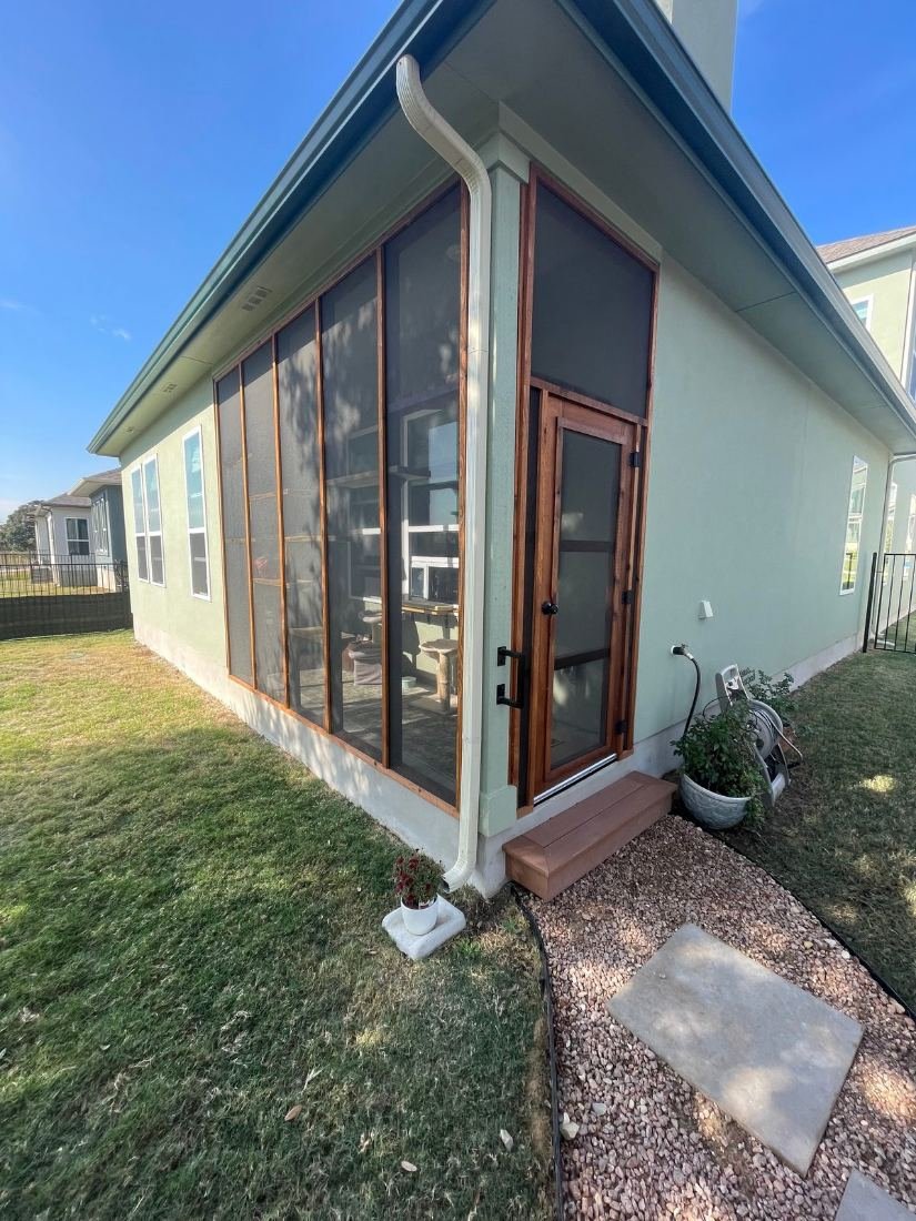 Corner of a screened porch catio on a sage green home, cedar-framed screen door with black hinges and a cedar step, climbing shelves and a cat tree visible inside
