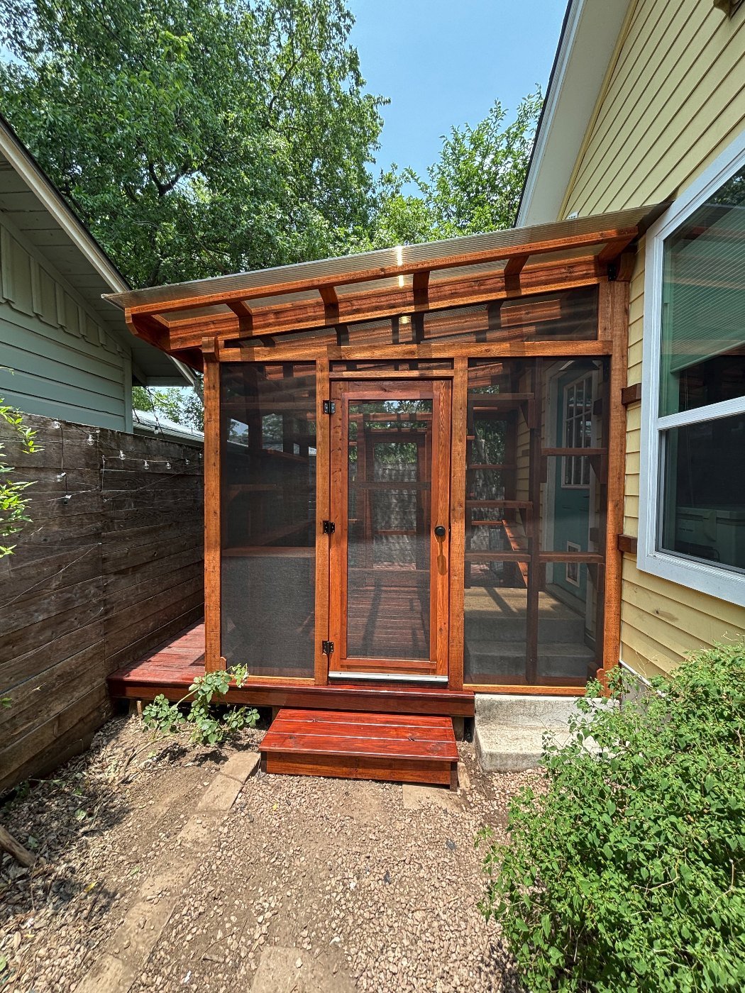 A cedar Signature catio with a bronze-tinted polycarbonate roof and a hand-built screen door, tucked beside a yellow bungalow