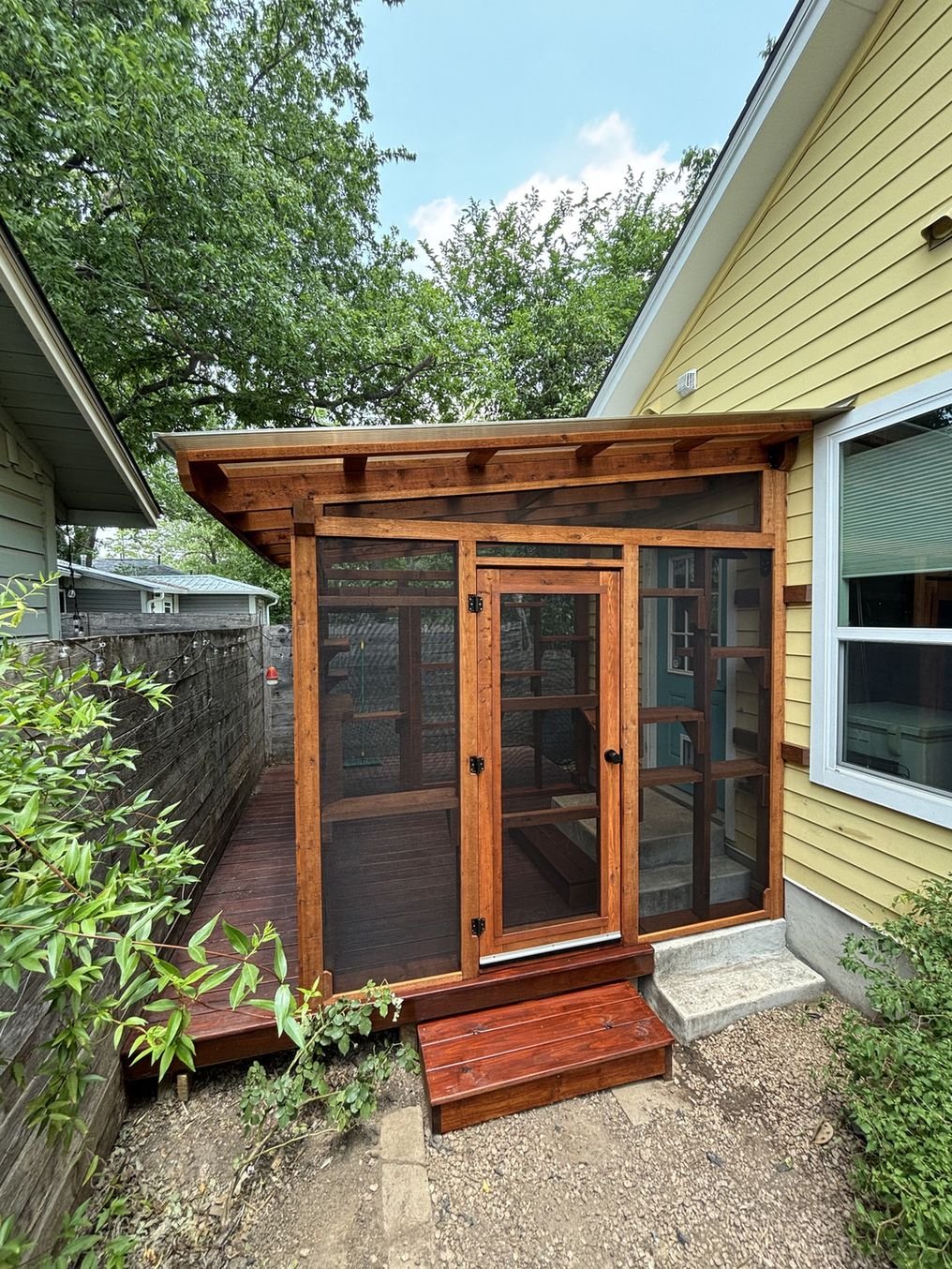 A tall cedar catio with a metal roof and handmade screen door, built on a deck beside a yellow bungalow