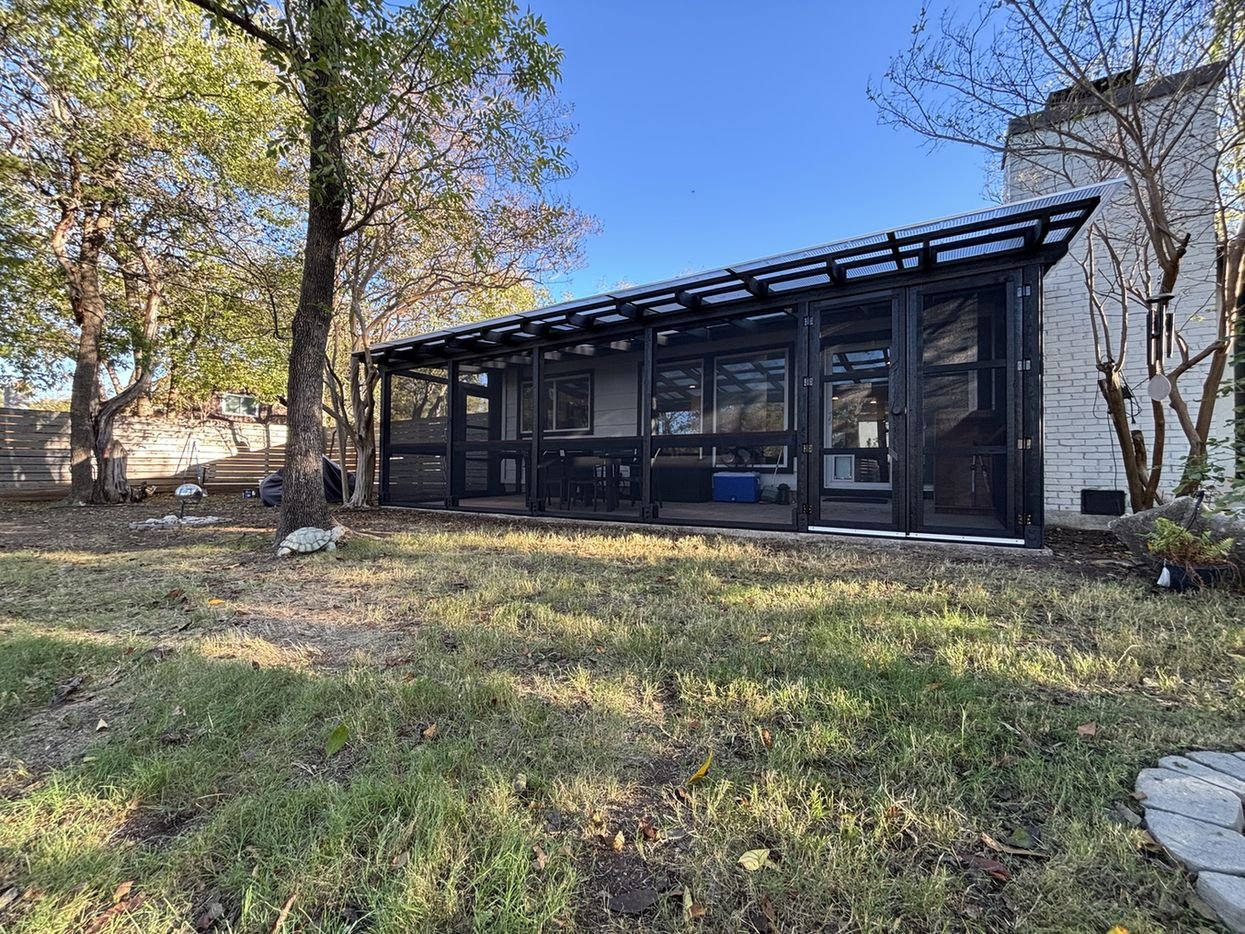 A long black-framed catio with a translucent roof, built between mature trees along a white brick house